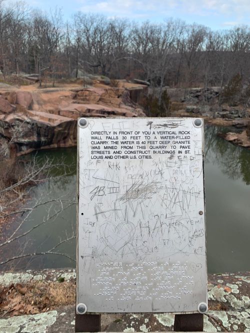 A weathered sign near a rocky area, detailing the history of the site and water depth, with visible graffiti.