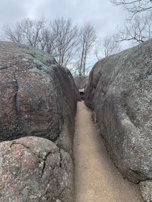 A narrow path between large, moss-covered boulders, surrounded by bare trees under a cloudy sky.