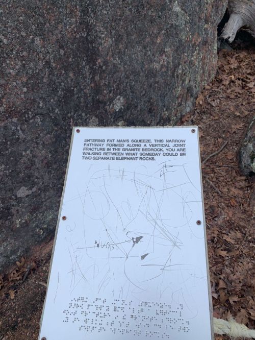 Sign at a rock formation, warning about a narrow passage called "Fat Man's Squeeze" with some scratches and markings.