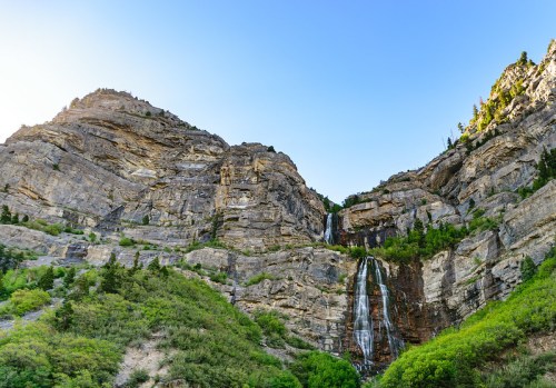 A stunning waterfall cascades down rocky cliffs, surrounded by lush greenery and a clear blue sky.