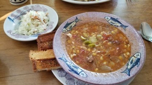 A bowl of hearty soup with vegetables and meat, served with coleslaw and two slices of toasted bread on a wooden table.