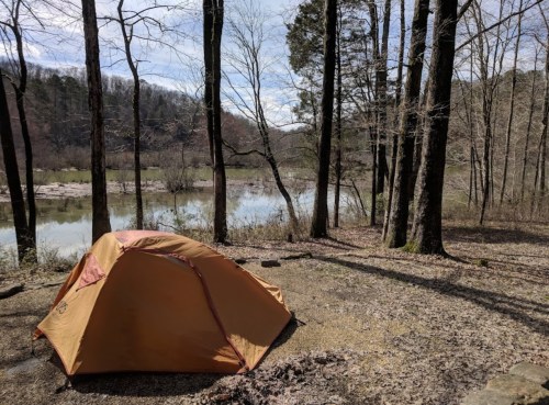 A bright orange tent set up near a calm river, surrounded by bare trees and a scenic landscape.