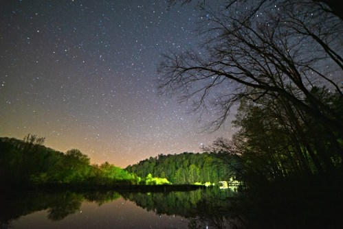 A serene night sky filled with stars reflects on a calm lake, surrounded by trees and distant hills.