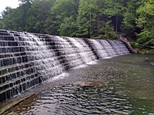 A cascading waterfall over a stone dam, surrounded by lush green trees and a calm river below.