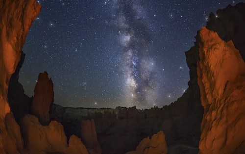 A stunning night sky filled with stars and the Milky Way, framed by rocky formations in a dark canyon.