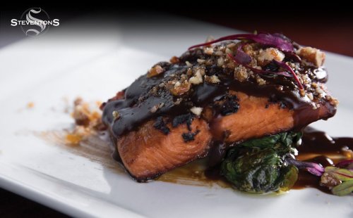 A beautifully plated salmon fillet topped with a rich sauce, nuts, and garnished with microgreens on a bed of spinach.