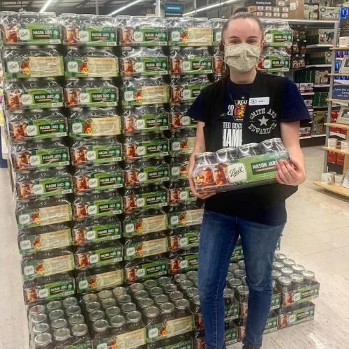 A person wearing a mask stands in a store aisle, holding mason jars in front of a display of stacked jars.