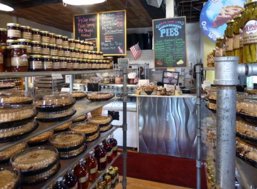 A cozy shop interior filled with jars of preserves and pies, featuring a chalkboard menu and a welcoming atmosphere.