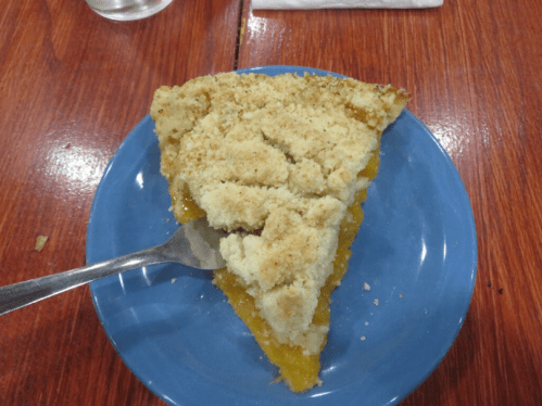 A slice of pie with a crumbly topping on a blue plate, resting on a wooden table. A fork is beside the plate.