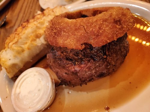 A plate featuring a juicy piece of meat with a crispy onion ring, served with a baked potato and dipping sauce.