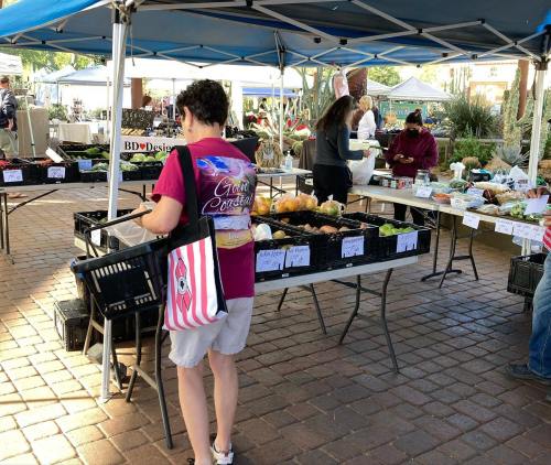 A woman shops at a farmers market, browsing fresh produce under blue tents with other shoppers in the background.