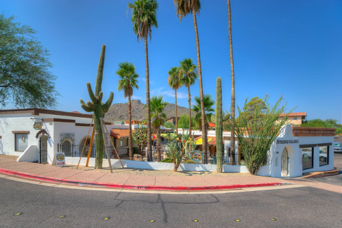 A sunny street view of a southwestern-style building surrounded by palm trees and cacti against a blue sky.