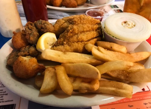 A plate of fried fish, hushpuppies, fries, and a slice of lemon, served with a side of onion and dipping sauce.