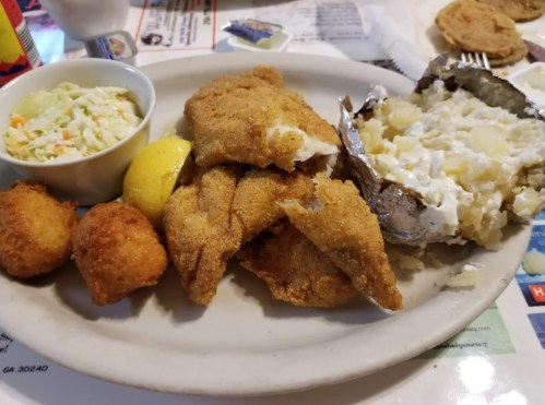 A plate of fried fish fillets, coleslaw, a baked potato with sour cream, and two hushpuppies.