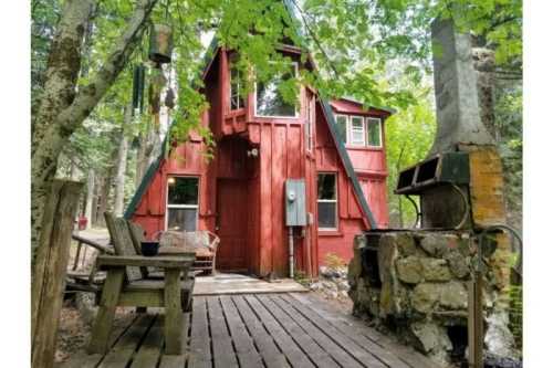 A red A-frame cabin surrounded by trees, with a wooden deck and stone fireplace in front.