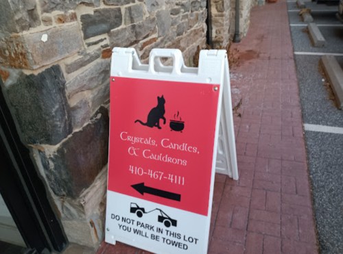 A sidewalk sign for "Rugs, Candles, & Cauldrons" featuring a cat graphic and parking instructions.