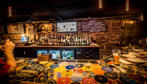 A cozy bar with a colorful record-themed countertop, shelves of liquor, and a chalkboard menu in a dimly lit setting.
