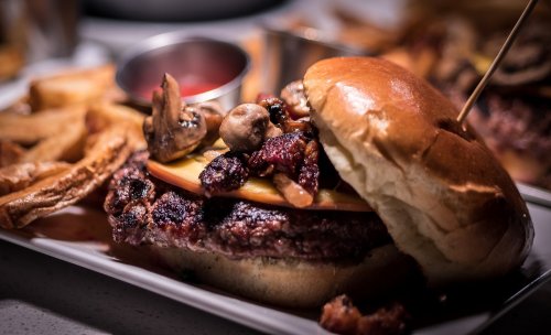 A gourmet burger topped with mushrooms and bacon, served with fries and a small cup of sauce on a white plate.