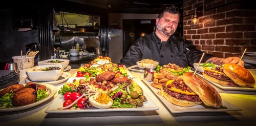 A chef stands behind a table filled with colorful dishes, including burgers, salads, and appetizers in a restaurant setting.