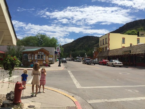 A sunny street scene with children walking, shops lining the road, and mountains in the background under a blue sky.