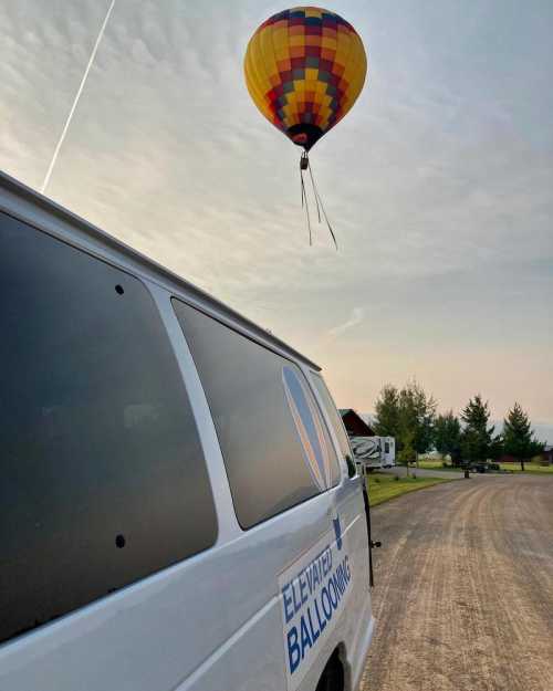 A colorful hot air balloon ascends into the sky above a gravel road, with a van labeled "Elevated Ballooning" in the foreground.