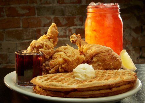 A plate of waffles topped with butter, fried chicken, a shot of syrup, and a drink in a jar.