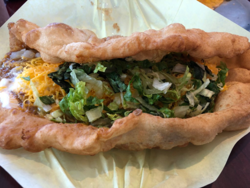A large, crispy taco filled with lettuce, cheese, and a savory meat mixture, served on a yellow paper plate.