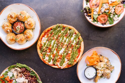A spread of dishes including pizza with pesto, stuffed bread, salad, and wings on a dark table.