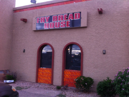 Exterior of Fry Bread House with a sign above arched windows and colorful wooden doors. Surrounding plants visible.