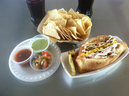 A hot dog with toppings, tortilla chips, and two dipping sauces on a tray, with drinks in the background.