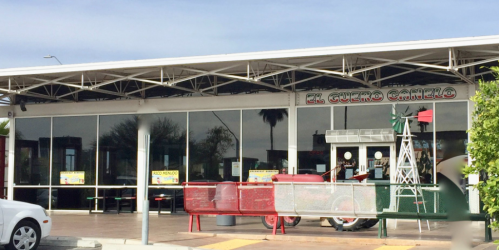 Exterior of a restaurant named "El Cuero Camelo," featuring large windows, a red tractor, and outdoor seating.