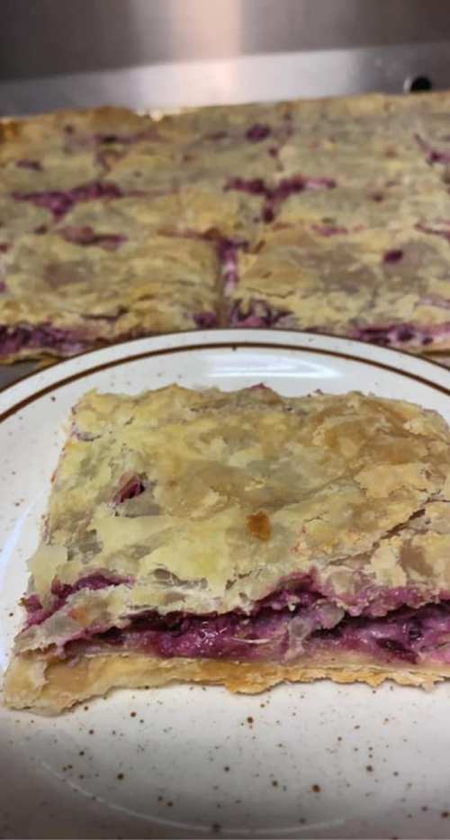 A slice of layered pastry with a purple filling, placed on a white plate, with more pastry in the background.