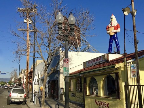 A street view featuring a colorful restaurant with a large chicken statue on the roof and utility poles in the background.