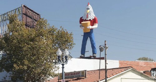 A large chicken statue wearing a red shirt and blue pants stands on a restaurant roof, holding a menu.