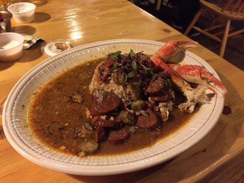 A plate of seafood gumbo with rice, sausage, okra, and crab claws, garnished with herbs on a wooden table.