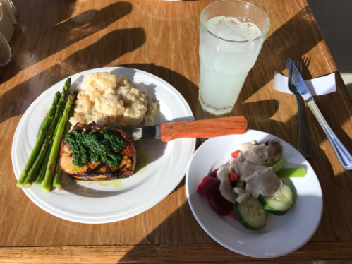 A plate with grilled salmon topped with green sauce, asparagus, mashed potatoes, and a side salad with dressing. A drink is beside it.