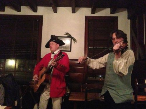 Two musicians perform in a cozy setting; one plays guitar in a red coat, while the other plays violin in a green vest.