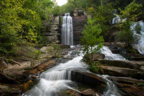 A serene waterfall cascades over rocky cliffs, surrounded by lush green trees and flowing water.