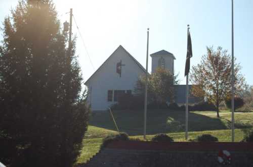 A white church building with a cross, surrounded by trees and flags, on a sunny day.