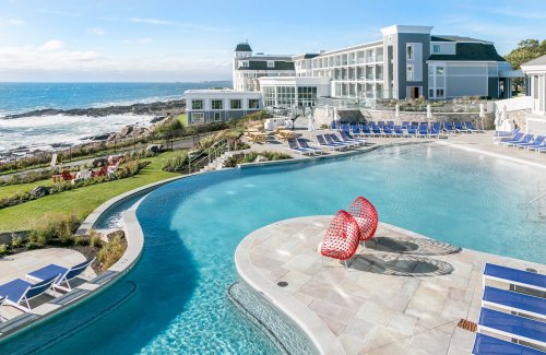 A scenic pool area with red lounge chairs, overlooking the ocean and a hotel in the background.