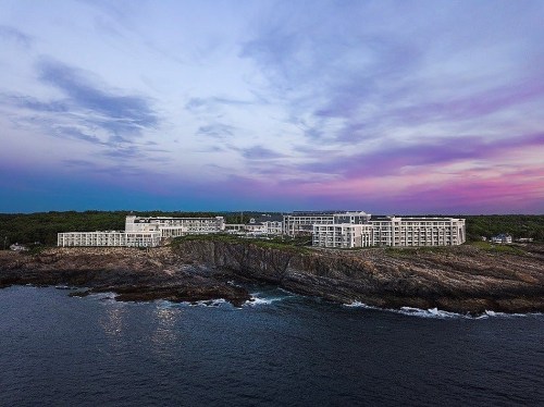 A modern hotel perched on a rocky coastline, with a colorful sunset sky and ocean waves below.