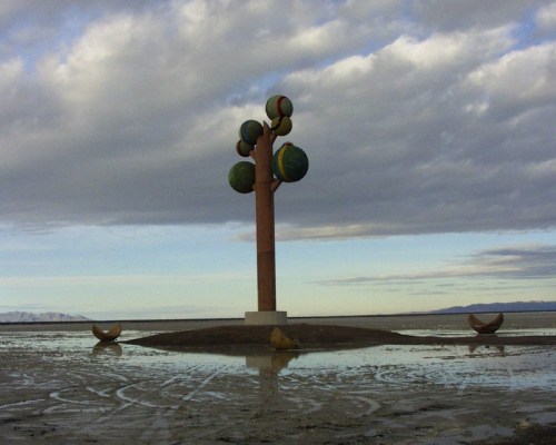 A colorful tree sculpture stands on a small island, surrounded by water and under a cloudy sky.