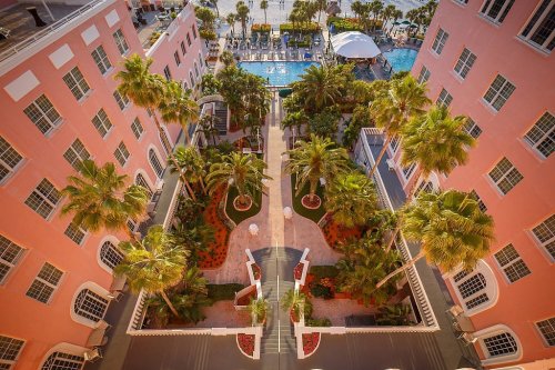 Aerial view of a pink hotel with palm trees and a pool area, surrounded by landscaped gardens.