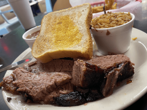 A plate with sliced brisket, a slice of buttered toast, rice, and a cup of beans on a table.