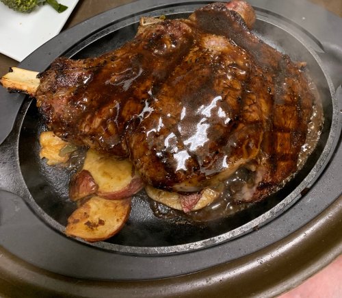 A sizzling platter of grilled meat with a glossy glaze, served on a bed of roasted potatoes. Steam rises from the dish.