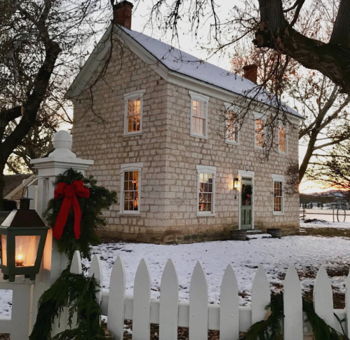 A charming stone house with a white picket fence, decorated for the holidays, surrounded by snow and trees at sunset.