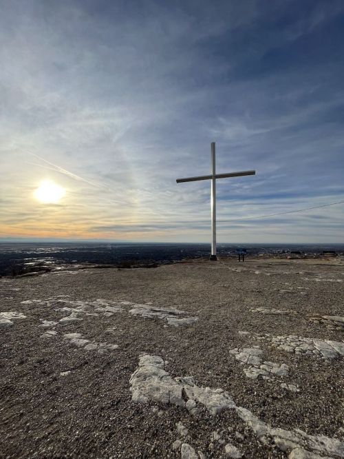 A large white cross stands on a rocky hilltop with a sunset in the background and two people in the distance.
