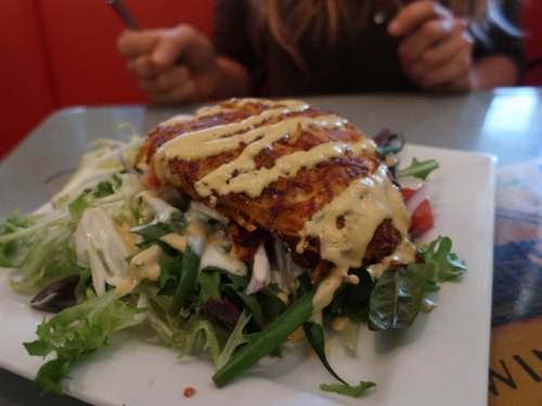 A plate of grilled chicken on a bed of mixed greens, drizzled with creamy sauce. Hands holding utensils in the background.