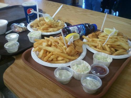 Two trays of food featuring fried fish, French fries, coleslaw, dipping sauces, and drinks on a wooden table.