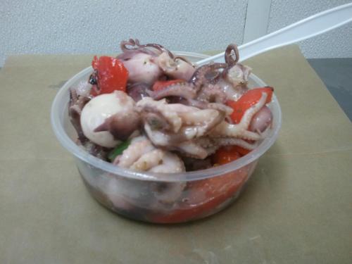 A clear plastic container filled with octopus, tomatoes, and other vegetables, served with a plastic spoon.